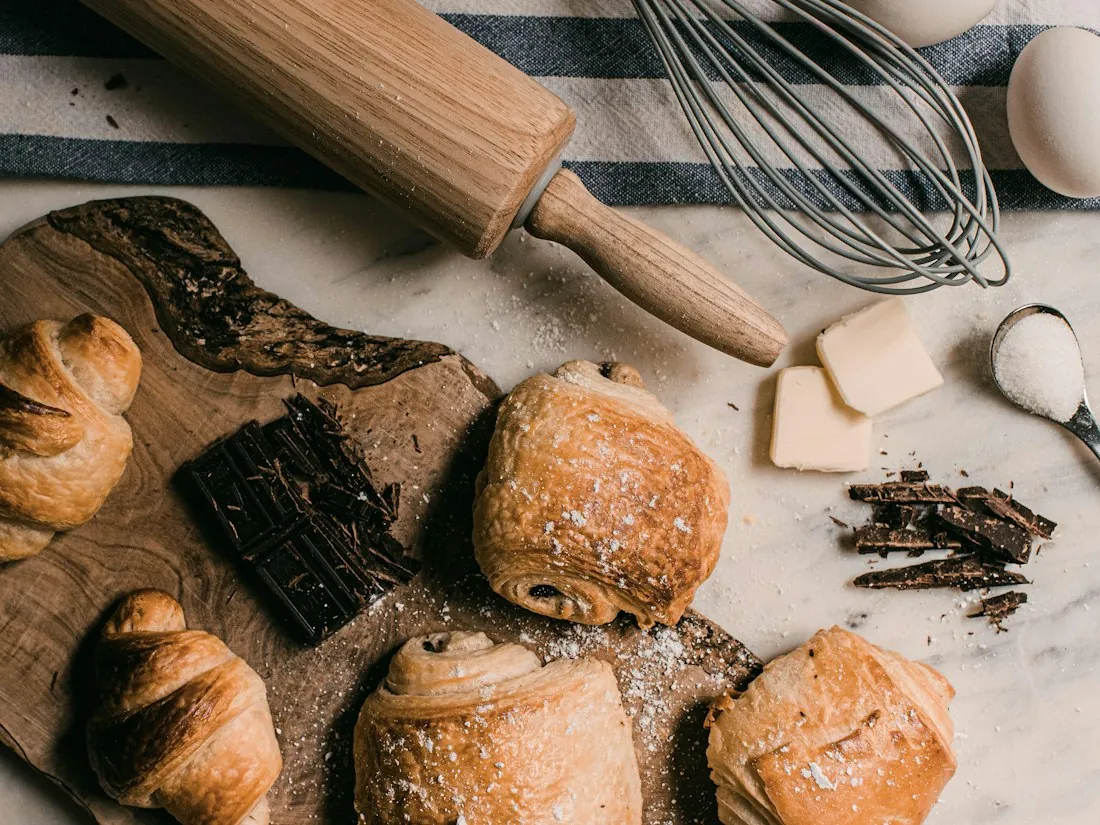 Croissants y pan de chocolate sobre una mesa de madera con rodillo y batidor